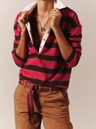 A person wearing a red and black horizontally striped knit polo shirt with a white collar, paired with brown pants and a patterned belt, against a plain beige studio background.