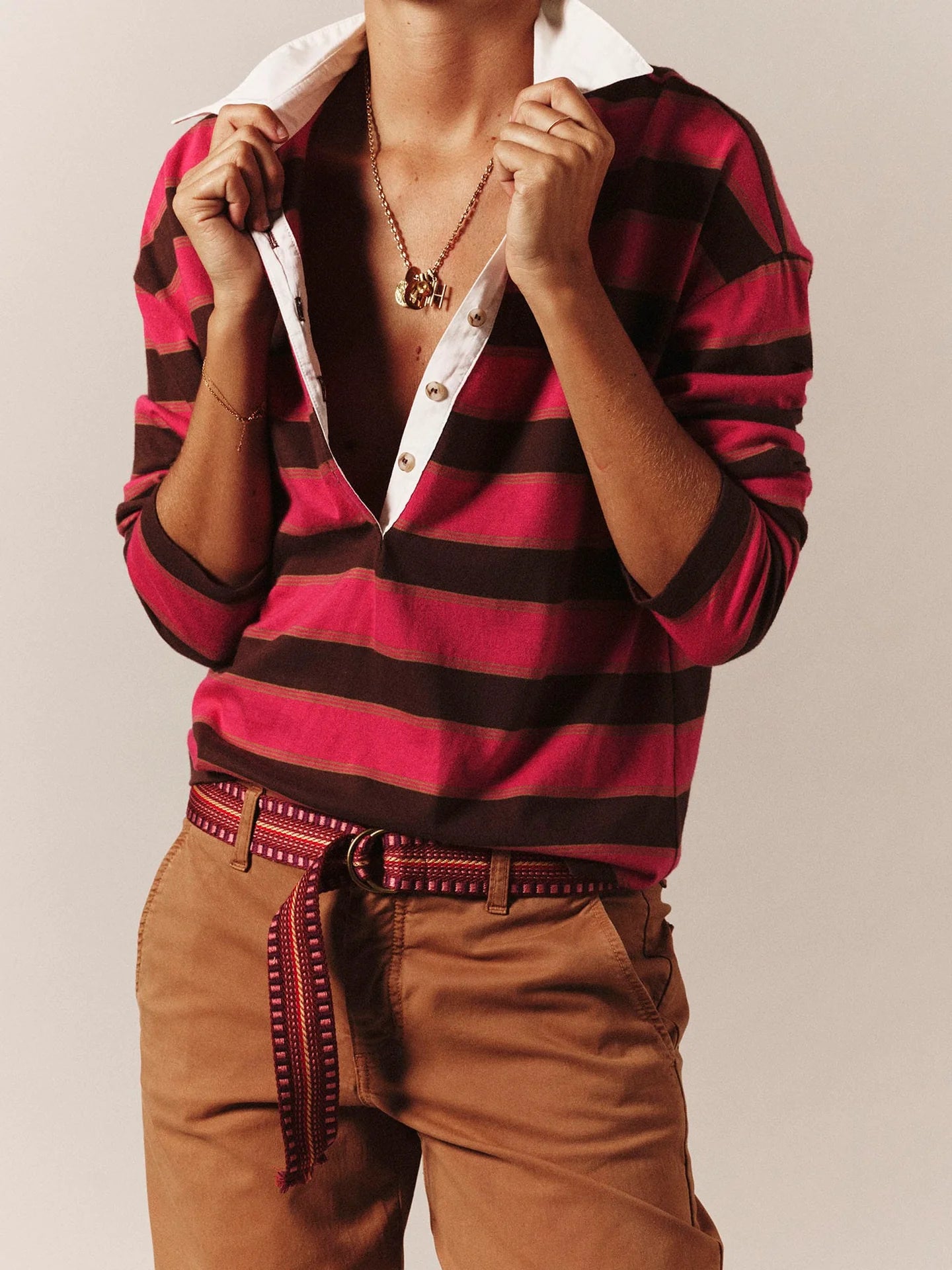 A person wearing a red and black horizontally striped knit polo shirt with a white collar, paired with brown pants and a patterned belt, against a plain beige studio background.
