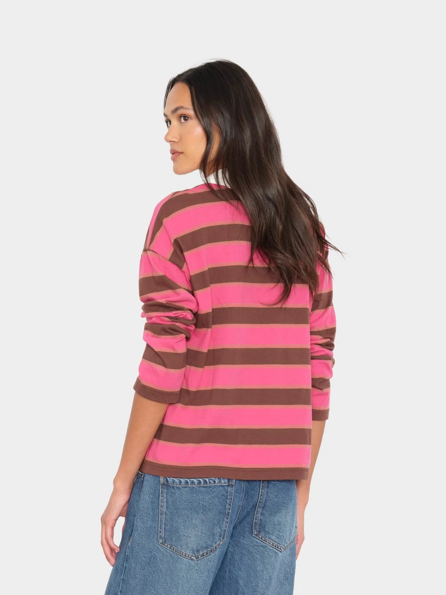 A woman shown from the back wearing a pink and brown horizontally striped long-sleeve top with blue jeans, against a light gray studio background.