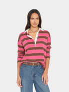 A woman modeling a pink and brown horizontally striped long-sleeve polo shirt with a white collar and button placket, paired with blue jeans, against a light gray background.