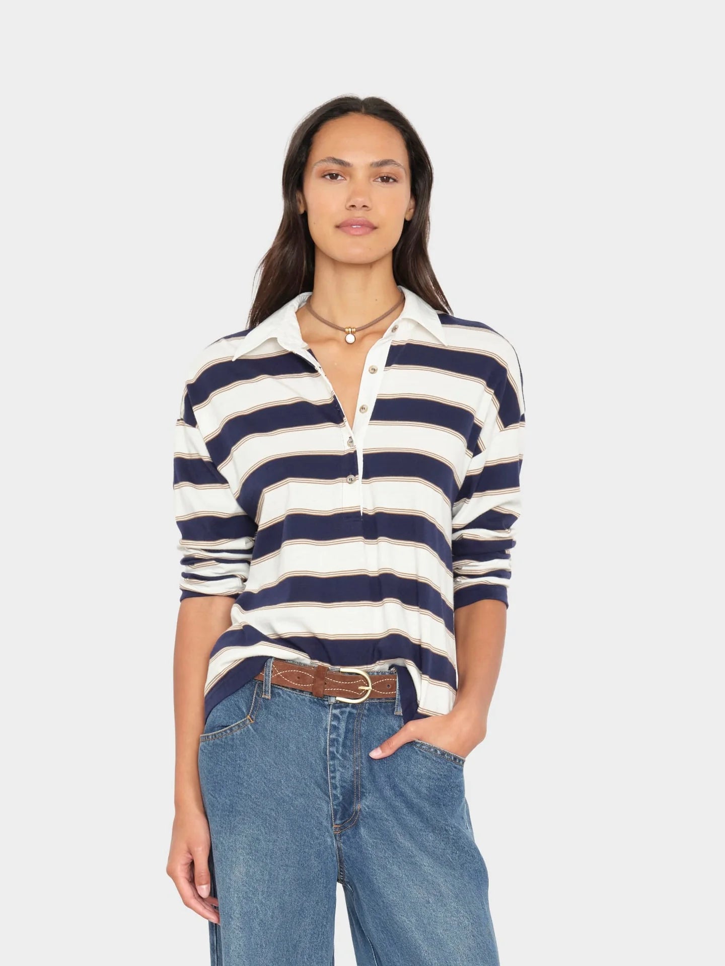 A woman modeling a navy and off-white horizontally striped knit polo shirt with a half-zip front, paired with blue jeans and a brown belt, against a light gray studio background.