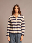 A woman modeling a long-sleeve collared knit top with horizontal navy and cream stripes, paired with blue jeans, against a solid taupe studio background.