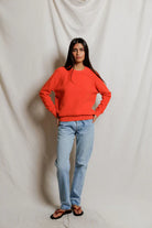 Woman wearing a bright orange sweater and light blue jeans against a plain background