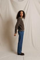 A woman stands in profile wearing a dark brown long-sleeve knit top, blue jeans, and dark brown shoes, posed against a draped off-white fabric backdrop.