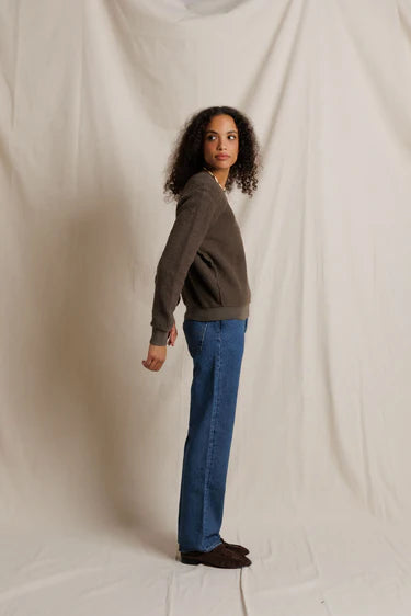 A woman stands in profile wearing a dark brown long-sleeve knit top, blue jeans, and dark brown shoes, posed against a draped off-white fabric backdrop.
