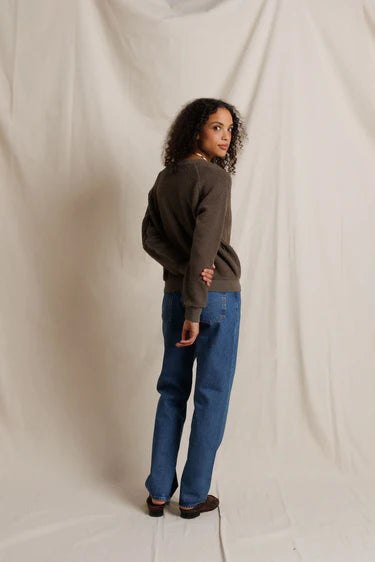 Person wearing a brown sweater and blue jeans standing against a plain background