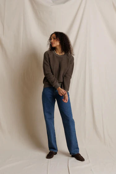 A woman modeling a dark brown crewneck sweater and blue jeans against a draped off-white fabric backdrop.
