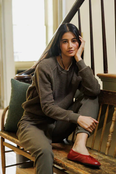 A woman sits on a wooden bench in a home interior, wearing a brown textured crewneck sweatshirt, olive green pants, and a red slip-on shoe. She is posed next to a green pillow and a dark wooden staircase.
