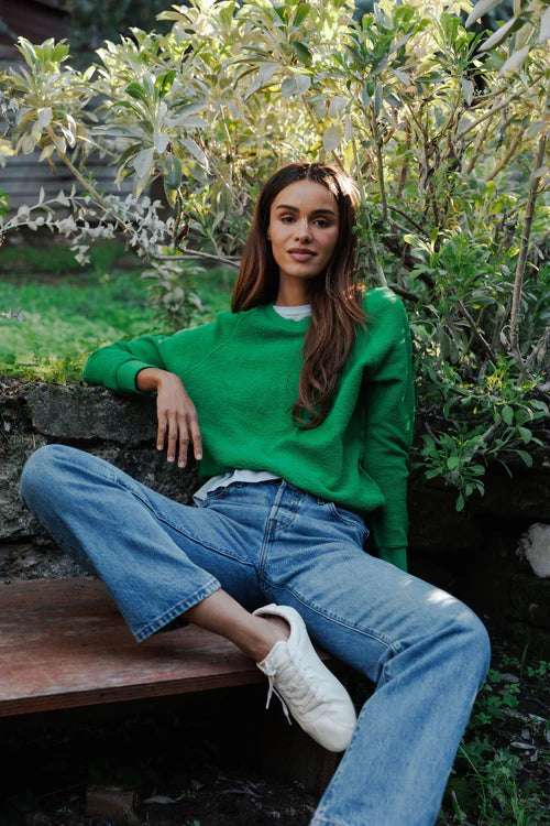 Woman in a green sweater and blue jeans sitting outdoors among plants