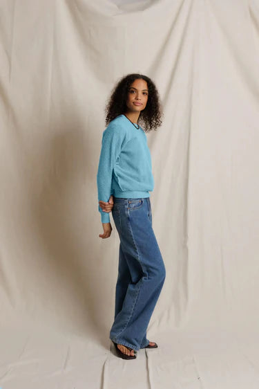 A woman modeling a light blue long-sleeve knit sweater and wide-leg blue jeans against a draped off-white fabric backdrop.