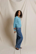 A woman modeling a light blue long-sleeve knit sweater and wide-leg blue jeans against a draped off-white fabric backdrop.
