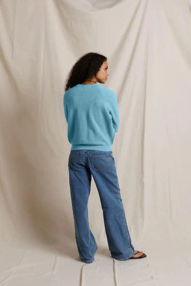 A woman stands with her back to the camera wearing a bright turquoise blue knit sweater and wide-leg blue jeans, posed against a draped off-white fabric backdrop.