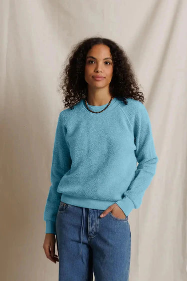 A woman with curly dark hair wearing a light blue crewneck sweater and blue jeans, standing against a draped beige fabric backdrop.
