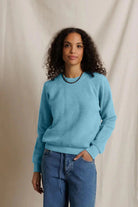 A woman with curly dark hair wearing a light blue crewneck sweater and blue jeans, standing against a draped beige fabric backdrop.