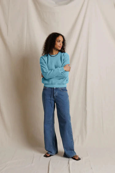 A woman stands against a draped cream fabric backdrop wearing a light blue crewneck sweatshirt, wide-leg blue jeans, and dark flat sandals.