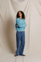 A woman stands against a draped off-white fabric backdrop, wearing a light blue crewneck sweatshirt and wide-leg blue jeans.