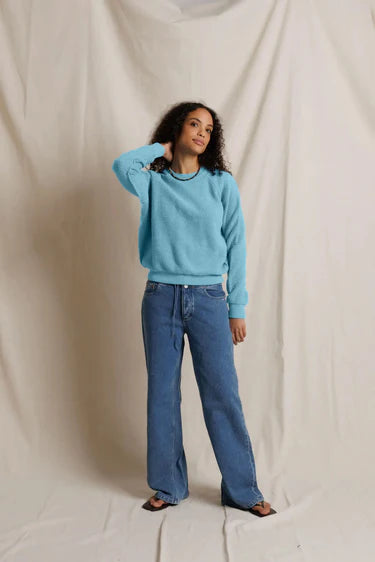 A woman modeling a light blue long-sleeve crewneck sweater and wide-leg blue jeans against a draped cream fabric backdrop.