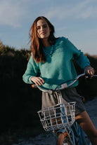Woman in a teal sweater riding a bicycle with a basket on a beach.