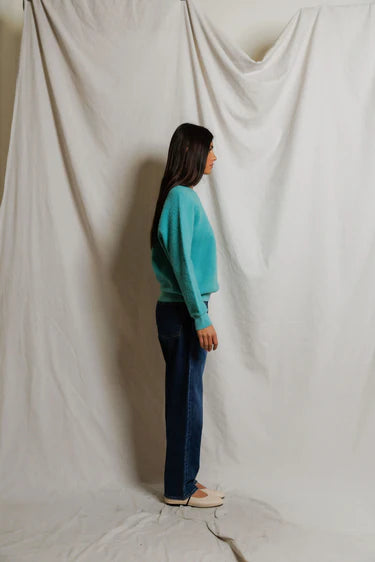 Person wearing a teal sweater and blue jeans standing against a white curtain.