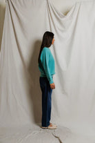 Person wearing a teal sweater and blue jeans standing against a white curtain.