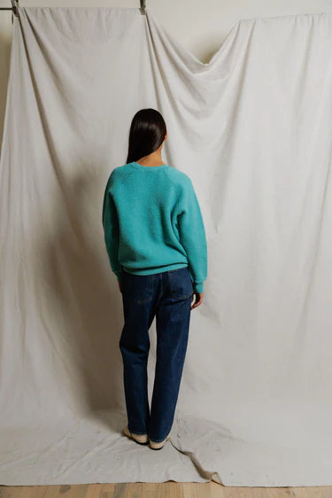 Person wearing a teal sweater and dark jeans standing against a plain white backdrop