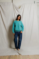 Person wearing a turquoise sweater and blue jeans standing against a plain white curtain.