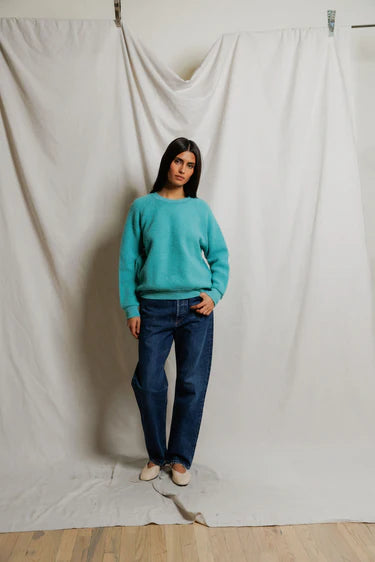 Person wearing a turquoise sweater and blue jeans standing against a plain white curtain.