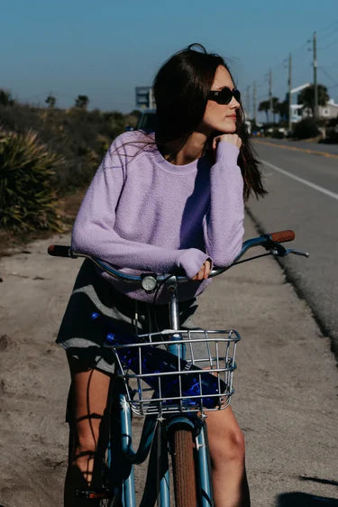 Woman in a purple sweater riding a bicycle on a road with a clear sky.