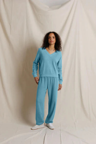 A woman stands against a draped cream fabric backdrop wearing a matching light blue knit lounge set consisting of a long-sleeve V-neck top and wide-leg pants, paired with white sneakers.