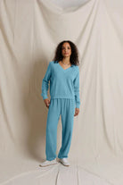 A woman stands against a draped cream fabric backdrop wearing a matching light blue knit lounge set consisting of a long-sleeve V-neck top and wide-leg pants, paired with white sneakers.
