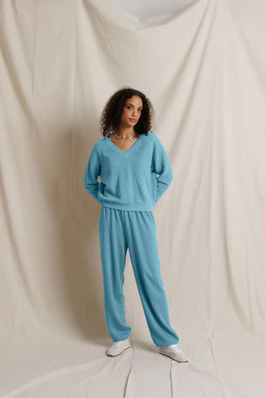A woman modeling a matching light blue lounge set consisting of a V-neck long-sleeve sweater and wide-leg sweatpants, paired with white sneakers, standing against a draped cream fabric backdrop.