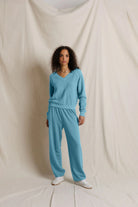 A model stands against a draped cream fabric backdrop wearing a matching light blue knit sweater and sweatpants set, with white sneakers.