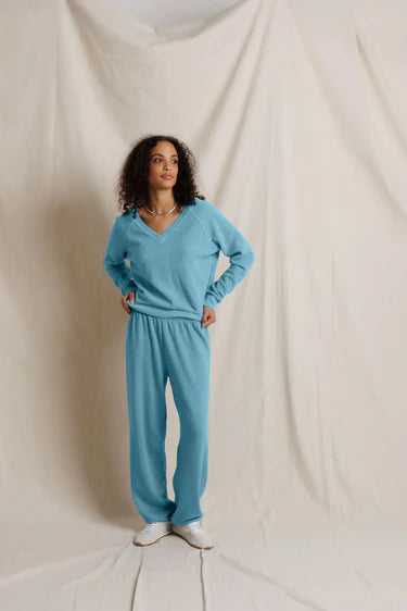 A woman modeling a matching light blue lounge set consisting of a long-sleeve V-neck top and wide-leg pants, standing against a draped off-white fabric backdrop.