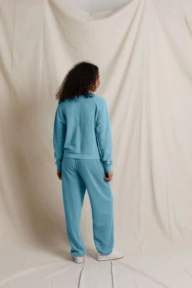 A person standing with their back to the camera, wearing a matching light blue sweatshirt and sweatpants set, against a draped cream fabric backdrop.