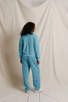 A person standing with their back to the camera, wearing a matching light blue sweatshirt and sweatpants set, against a draped cream fabric backdrop.